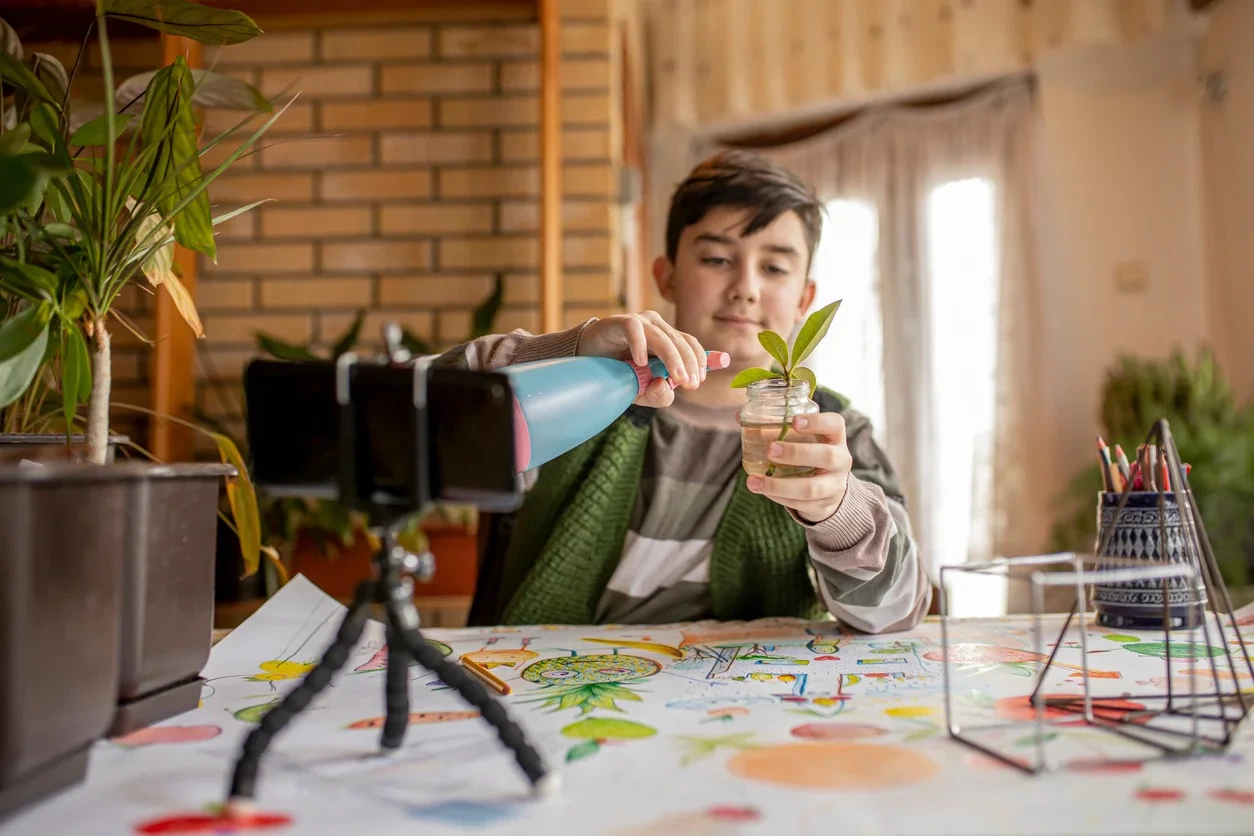 junger Schüler besprüht eine Pflanze mit Wasser und dokumentiert das mit einer Handykamera