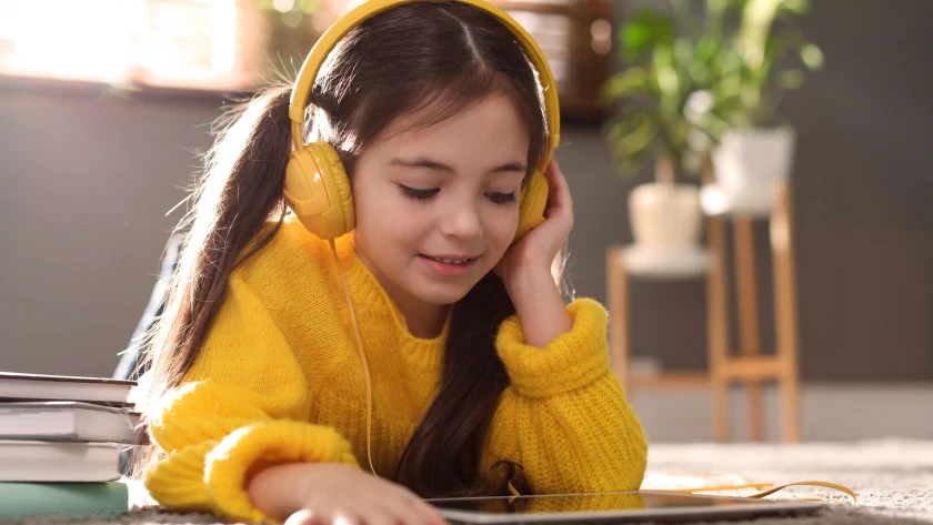 Mädchen mit gelbem Pulli liegt auf dem Boden und hört mit Kopfhörern etwas auf ihrem Tablet an
